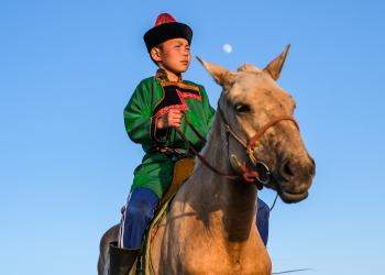 A young boy dressed in vibrant traditional attire riding a horse under a clear blue sky